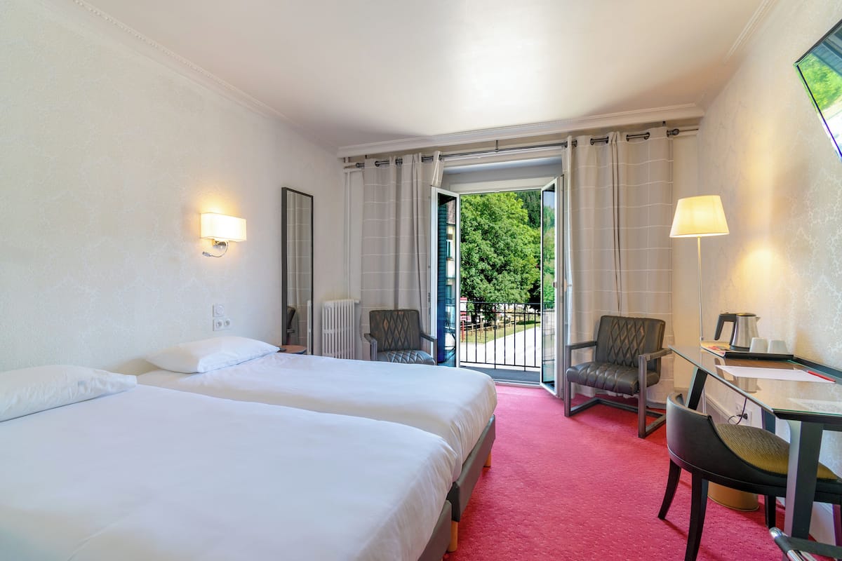 A hotel room with two beds, a desk, a chair, a lamp, and a view of greenery through the open door.