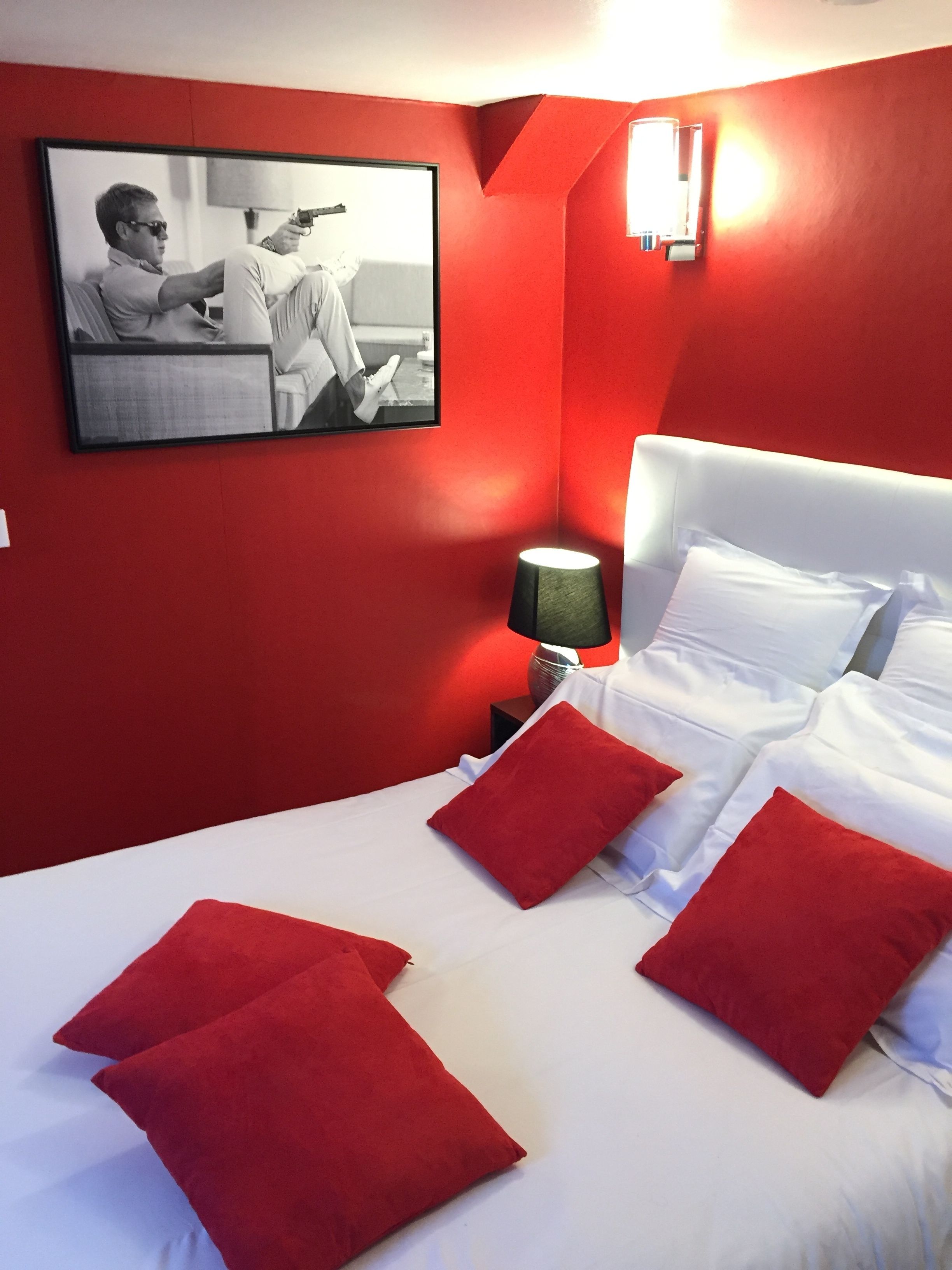 Ein Hotelzimmer mit roter Akzentwand, einem Bett mit weißer Bettwäsche und roten Kissen, einem Nachttisch mit Lampe und einem gerahmten Bild an der Wand.
