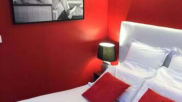 Ein Hotelzimmer mit roter Akzentwand, einem Bett mit weißer Bettwäsche und roten Kissen, einem Nachttisch mit Lampe und einem gerahmten Bild an der Wand.