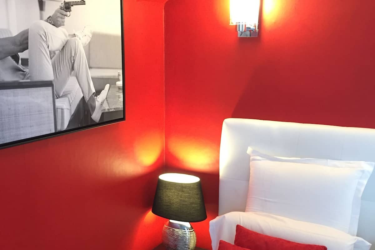 Hotellihuone, jossa on punainen seinä, valkoinen sänky punaisilla tyynyillä, yöpöytä lampun kanssa ja seinällä oleva kehystetty kuva.