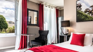 Hotellihuone, jossa on sänky, tuoli, pöytä, peili ja taulu seinällä.