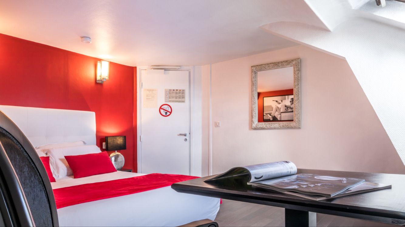 A hotel room with a bed, a red wall, a white door, a framed picture, and a table with magazines.