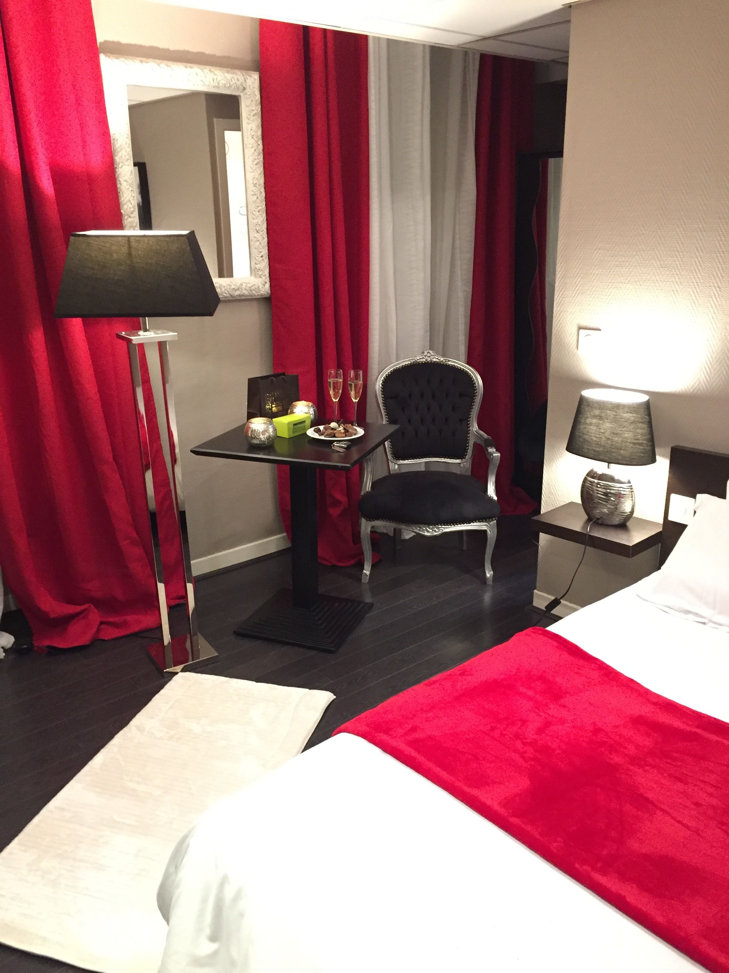Hotelkamer met een bed, een stoel, een tafel met fles en glazen, een lamp en rode gordijnen.