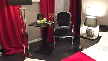 Hotelkamer met een bed, een stoel, een tafel met fles en glazen, een lamp en rode gordijnen.