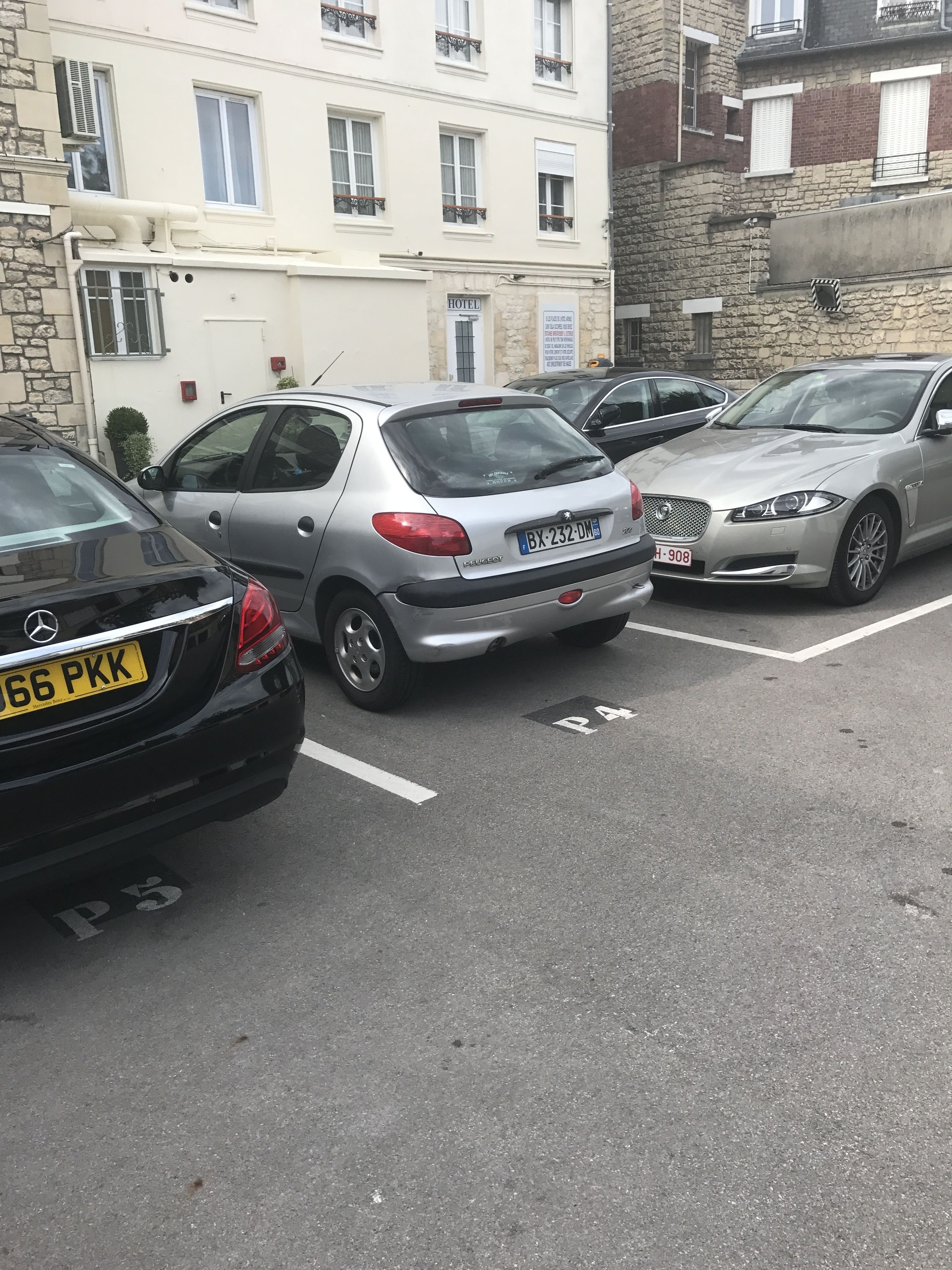 Un estacionamiento con varios coches, incluyendo un Mercedes-Benz plateado y un coche negro con matrícula amarilla.