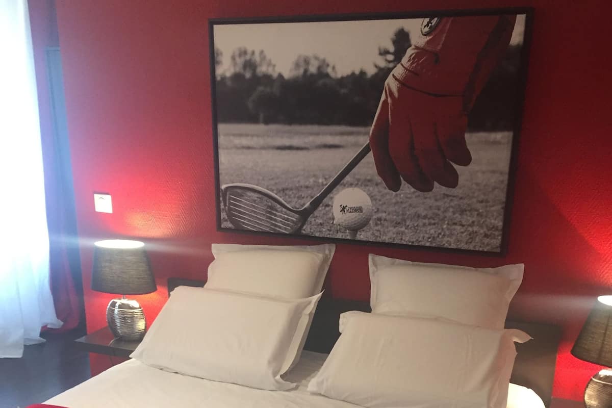 Hotellihuone, jossa on sänky, valkoiset vuodevaatteet, punainen heittopeitto ja seinällä kehystetty kuva golflyönnistä.