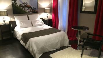 A neatly made bed with white linens, a small round table, and a chair, with a painting on the wall and a mirror above the bed.