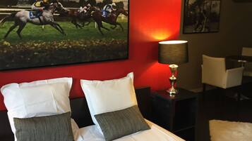 Une chambre d’hôtel avec un grand lit, deux oreillers, un mur d’accent rouge, un tableau représentant des jockeys, une lampe et une chaise.