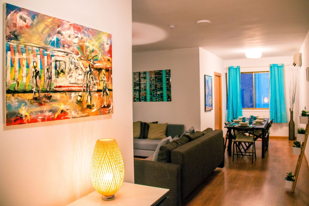 A living room with a sofa, a table with chairs, a painting, and a lamp.