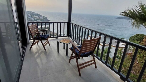 Un balcón con dos sillas de madera y una mesa pequeña, con vistas a la costa y un cielo despejado.