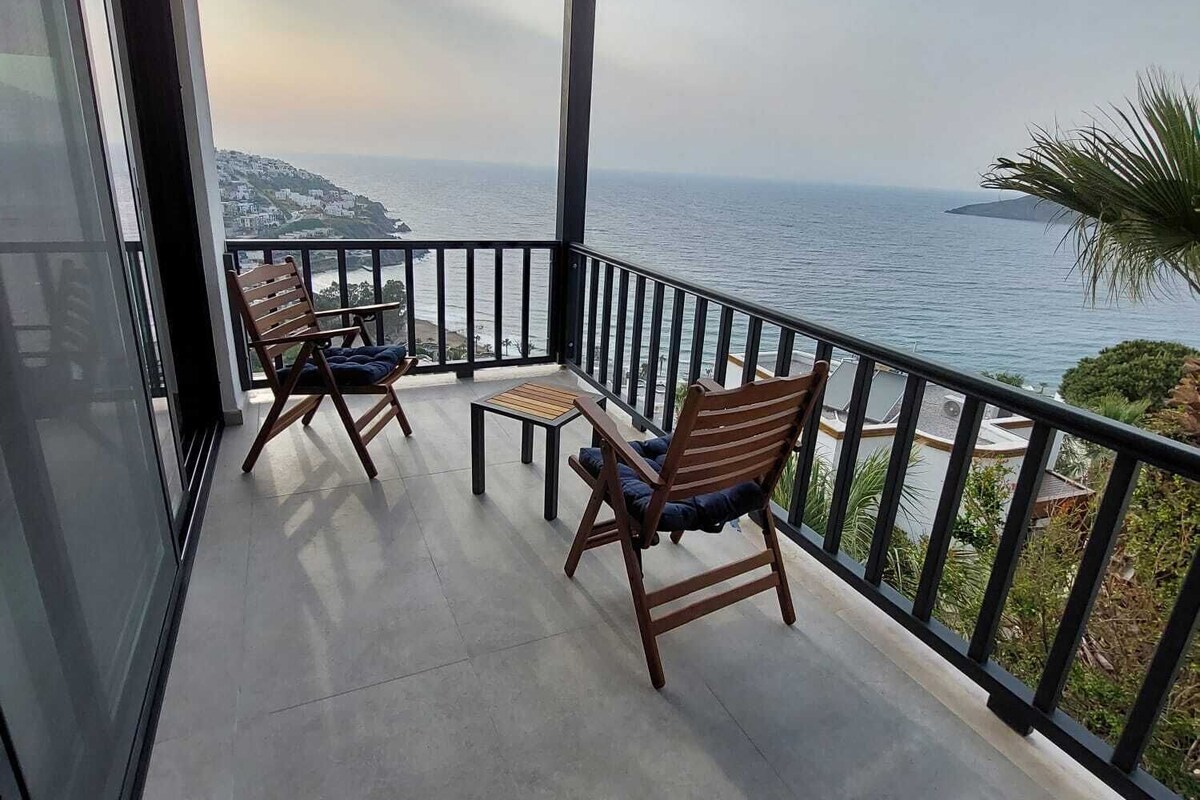 Un balcon avec deux chaises en bois et une petite table, offrant une vue sur la côte et un ciel dégagé.