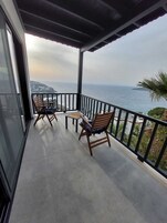 Varanda com duas cadeiras de madeira e uma mesa pequena, com vista para a costa e céu claro.
