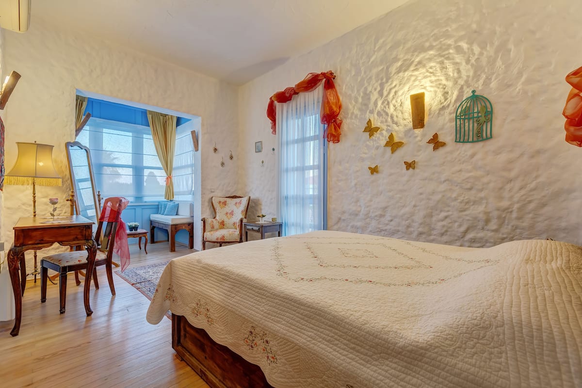 A bedroom with a large bed, a desk with a chair, a window with curtains, and decorative wall elements.