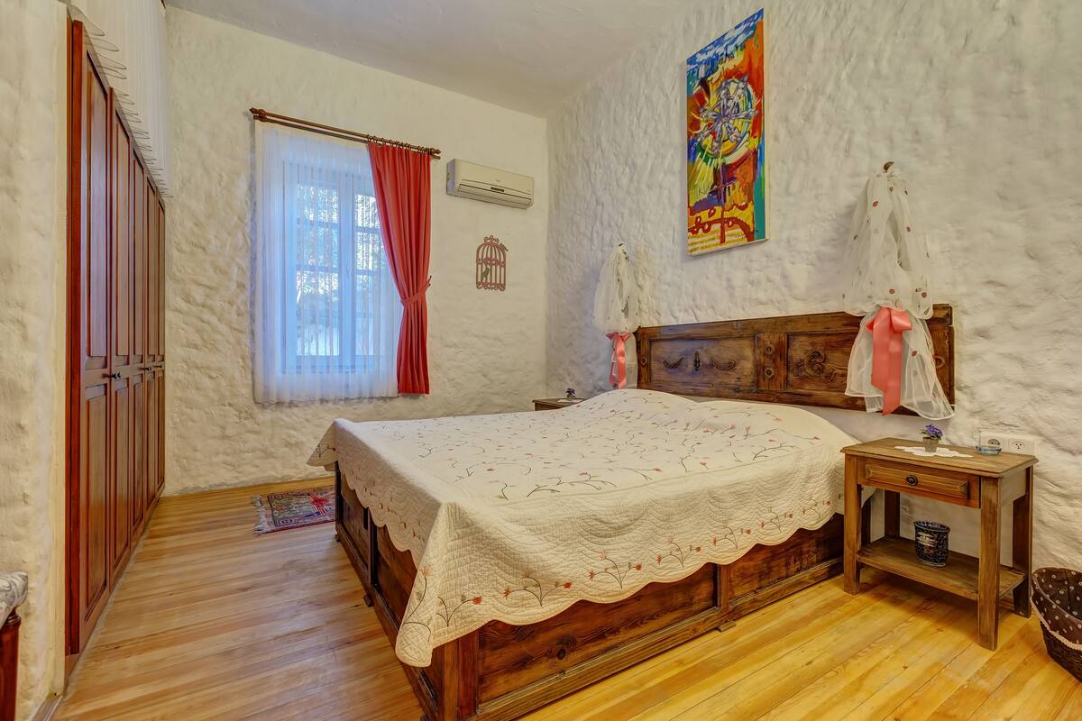A bedroom with a wooden bed, a nightstand, a window with curtains, and a wall-mounted air conditioner.