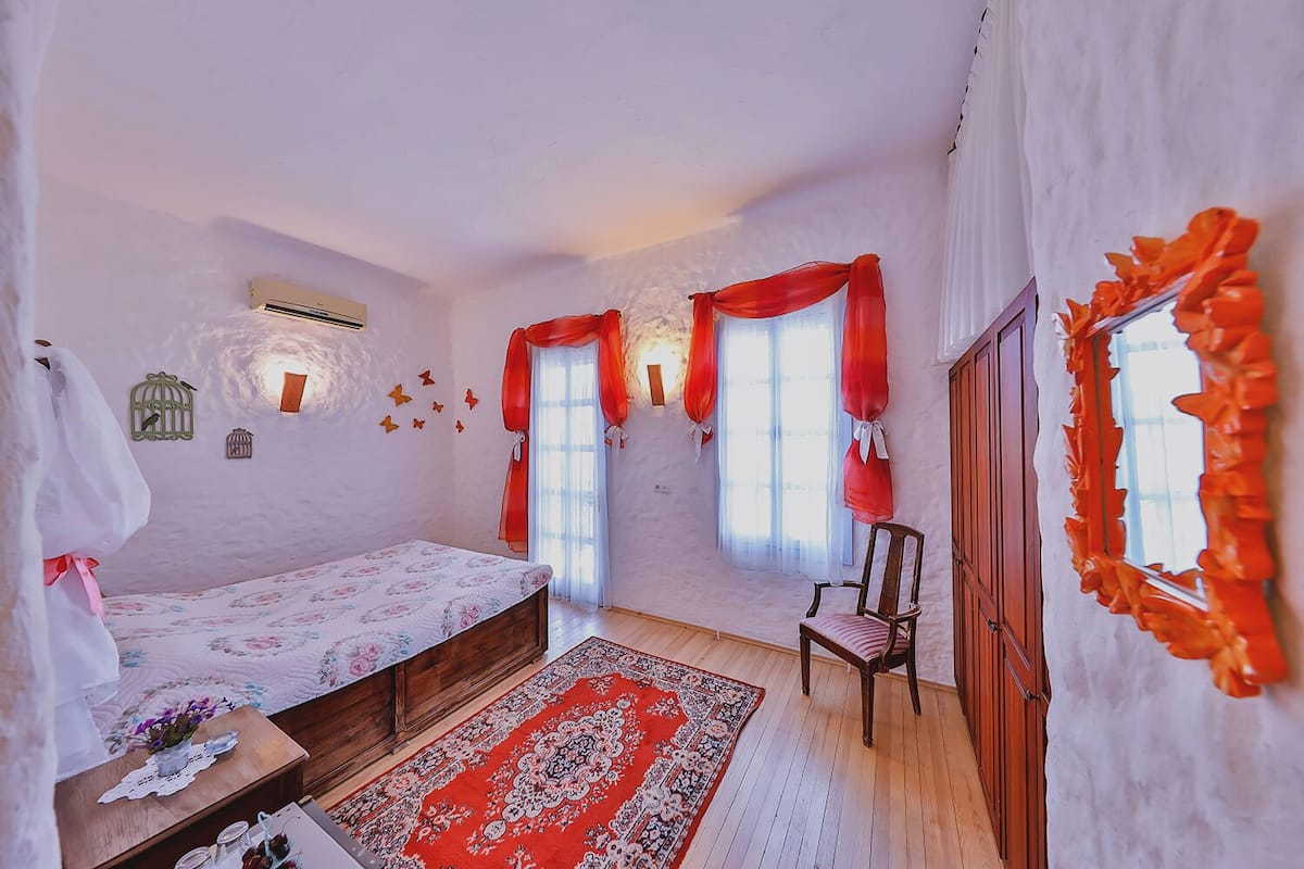 A bedroom with a bed, a chair, a window with curtains, a wall-mounted air conditioner, and a decorative mirror.