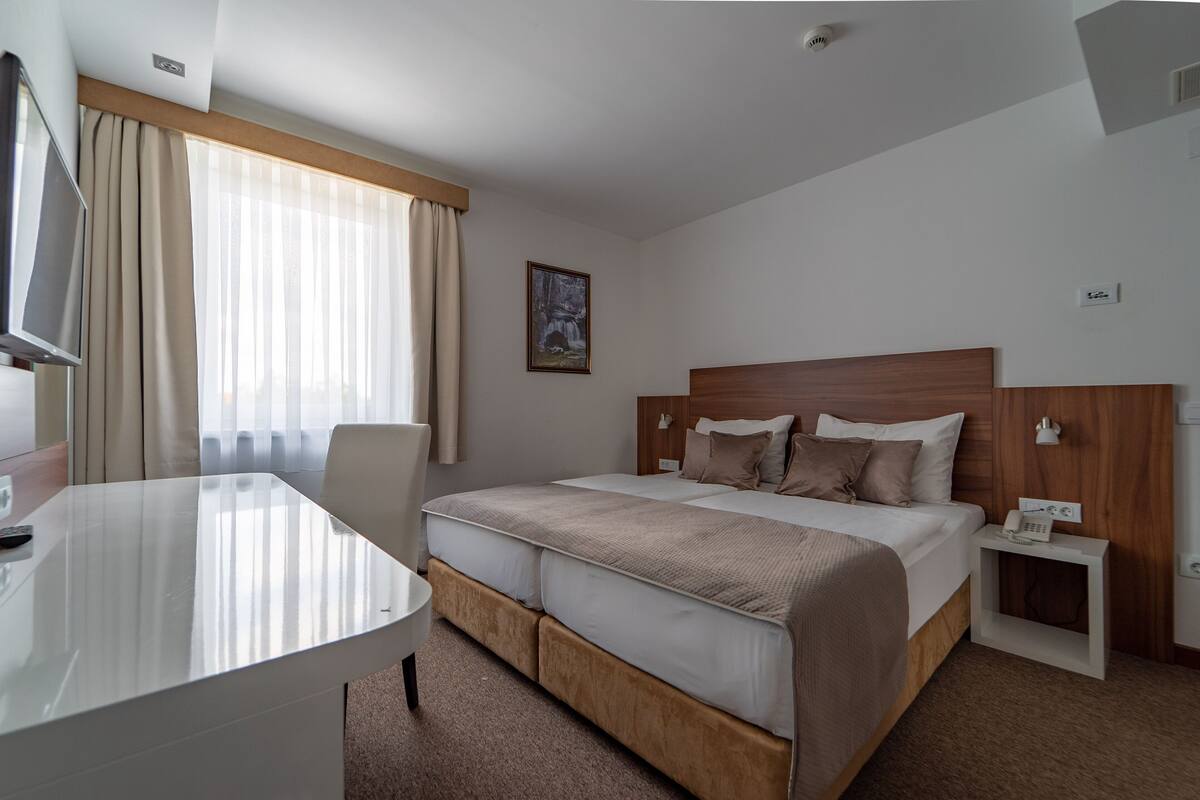 A hotel room with a bed, bedside table, chair, desk, and a painting on the wall.