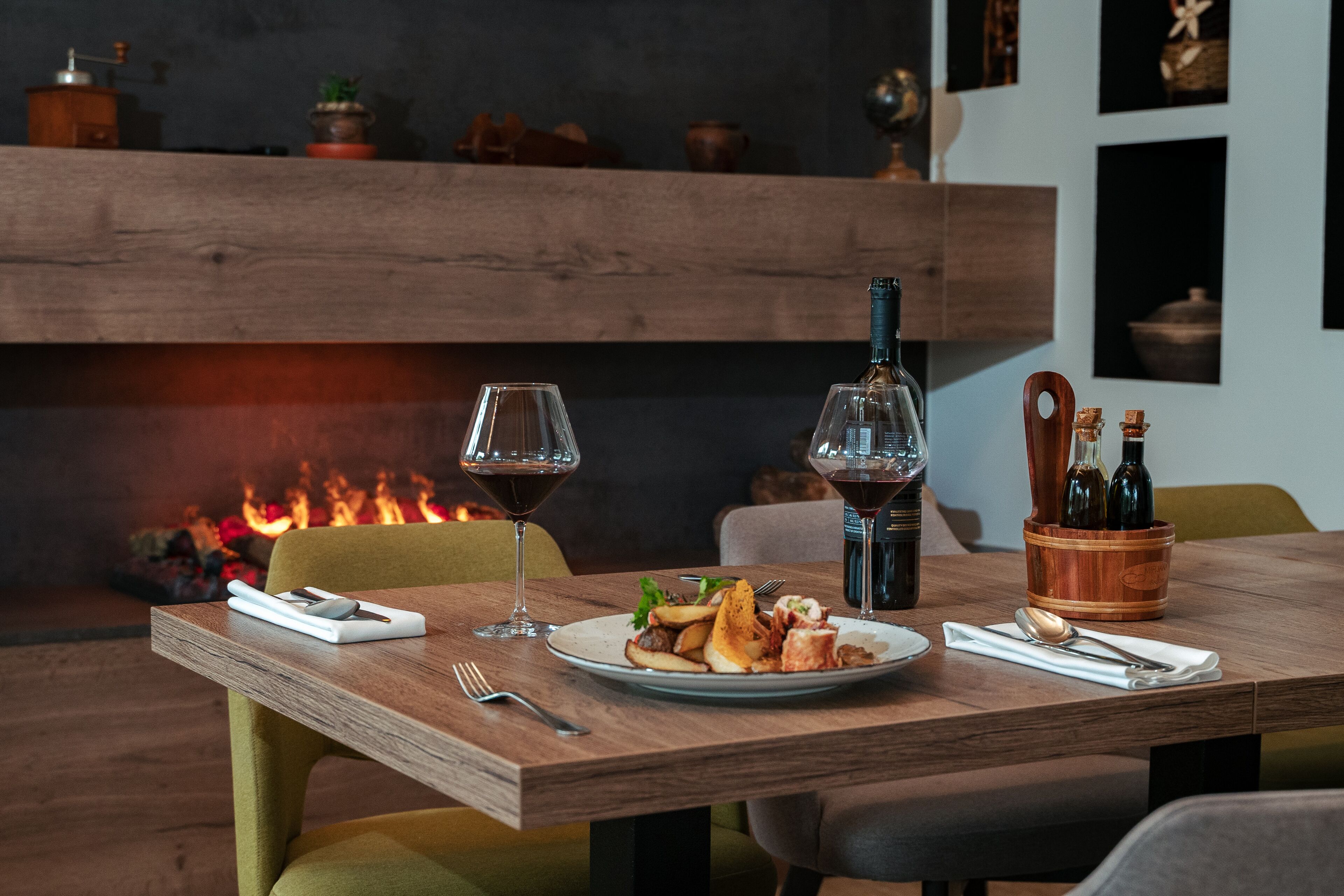 A wooden dining table with a plate of food, two wine glasses, a bottle, and a wooden bottle holder.
