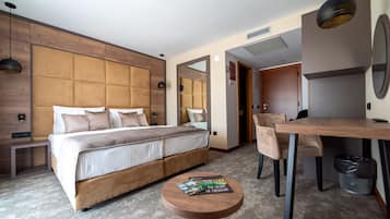 A modern hotel room with a large bed, a wooden headboard, a round table with a magazine, a chair, and a small trash bin.
