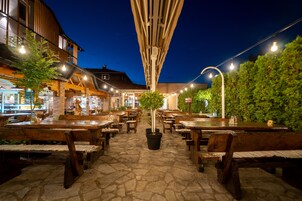 Un área de comedor al aire libre con suelo de piedra, mesas y bancos de madera, luces colgantes y vista a un edificio de fachada de ladrillo.
