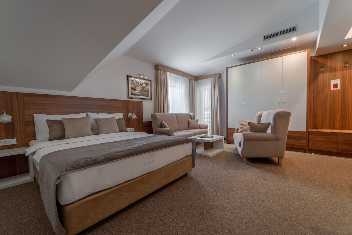 A modern hotel room with a large bed, two armchairs, a small table, and a wardrobe.