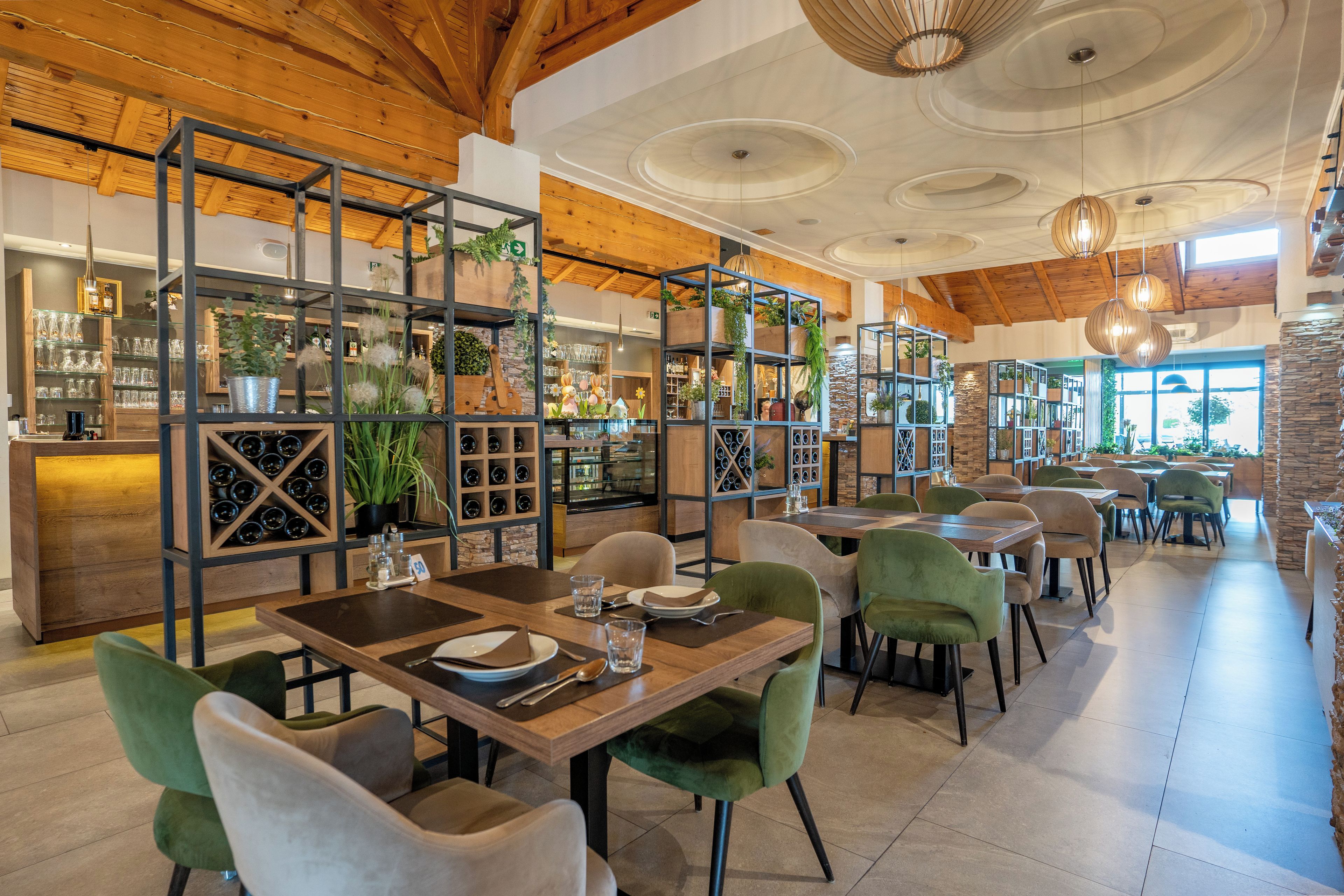 A restaurant interior with wooden beams, tiered shelving with plants, and green upholstered chairs.