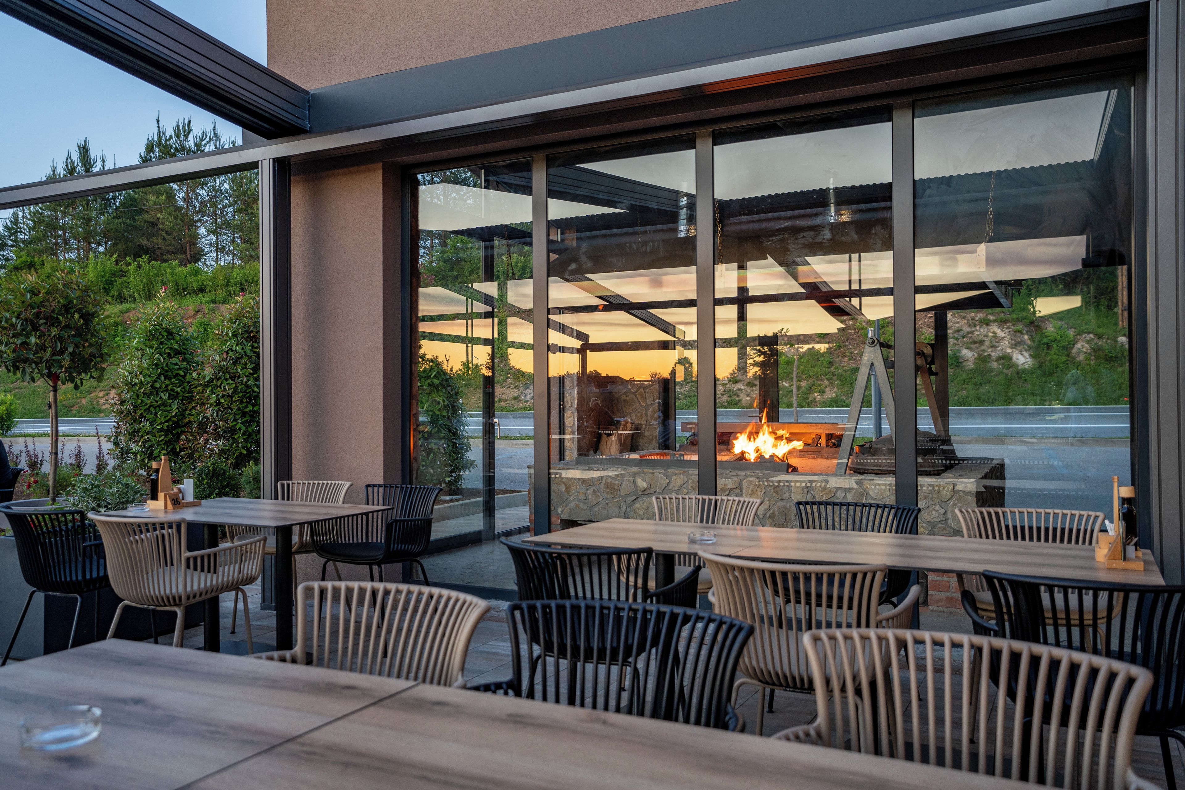 An outdoor seating area with tables and chairs, a fire feature, and a view through large windows.
