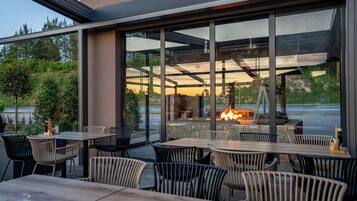 An outdoor seating area with tables and chairs, a fire feature, and a view through large windows.