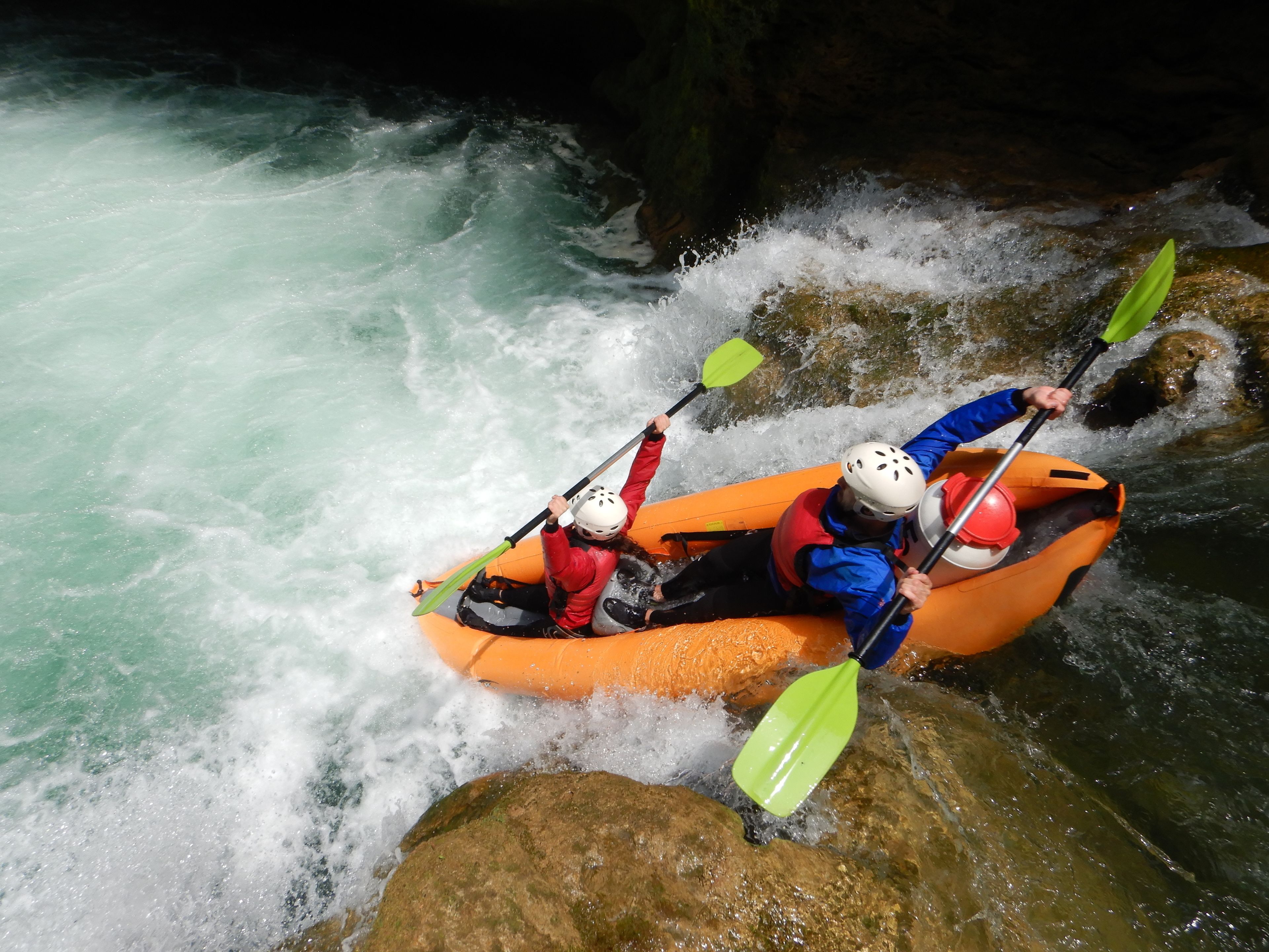 Two people kayaking in an orange raft on a river with white water.