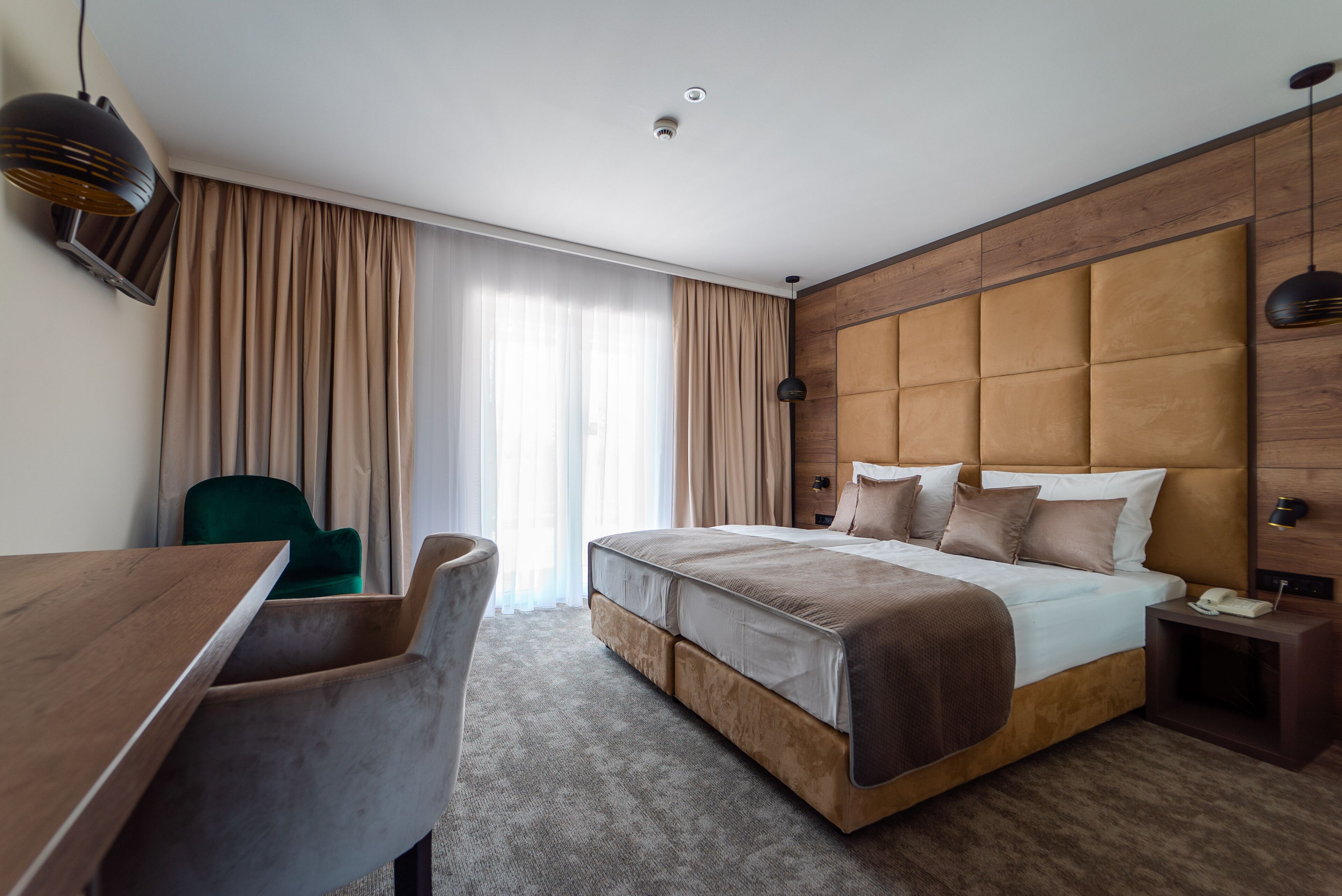 A hotel room with a large bed, a desk, and a chair. There is a window with curtains, a wooden headboard, and a nightstand with a lamp.