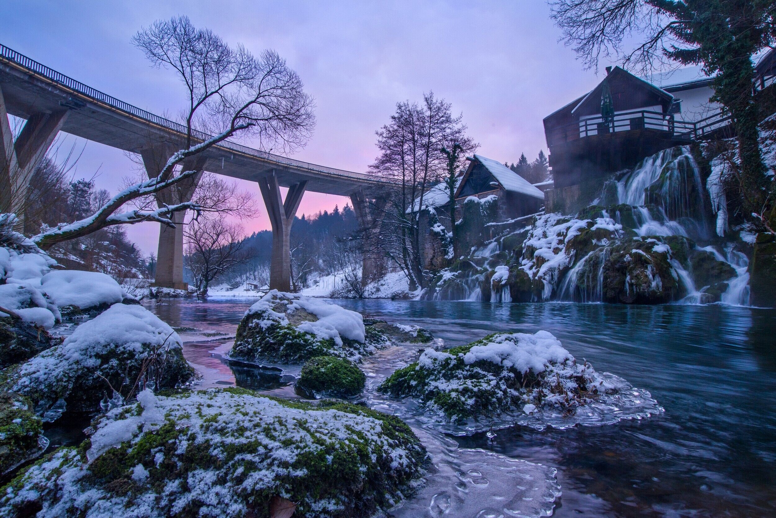 A winter scene with a bridge, a cabin, and a flowing river.