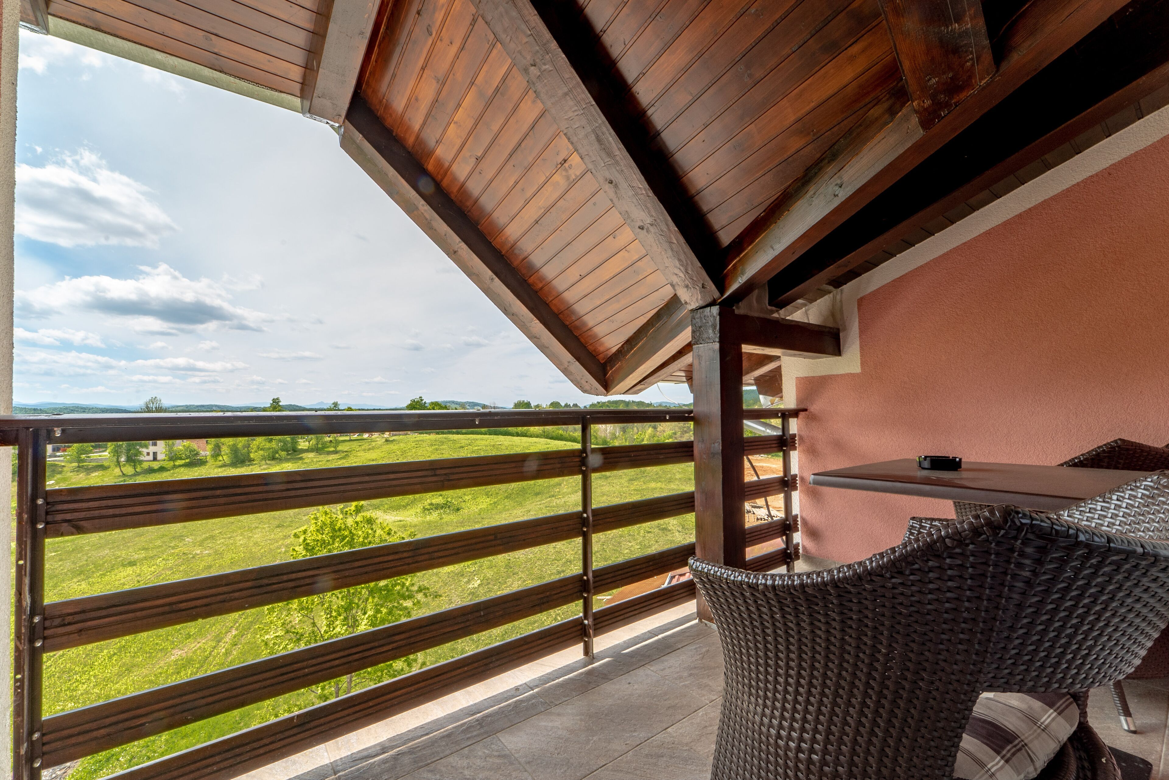 Un balcon donnant sur un paysage verdoyant, une chaise en osier et une table.