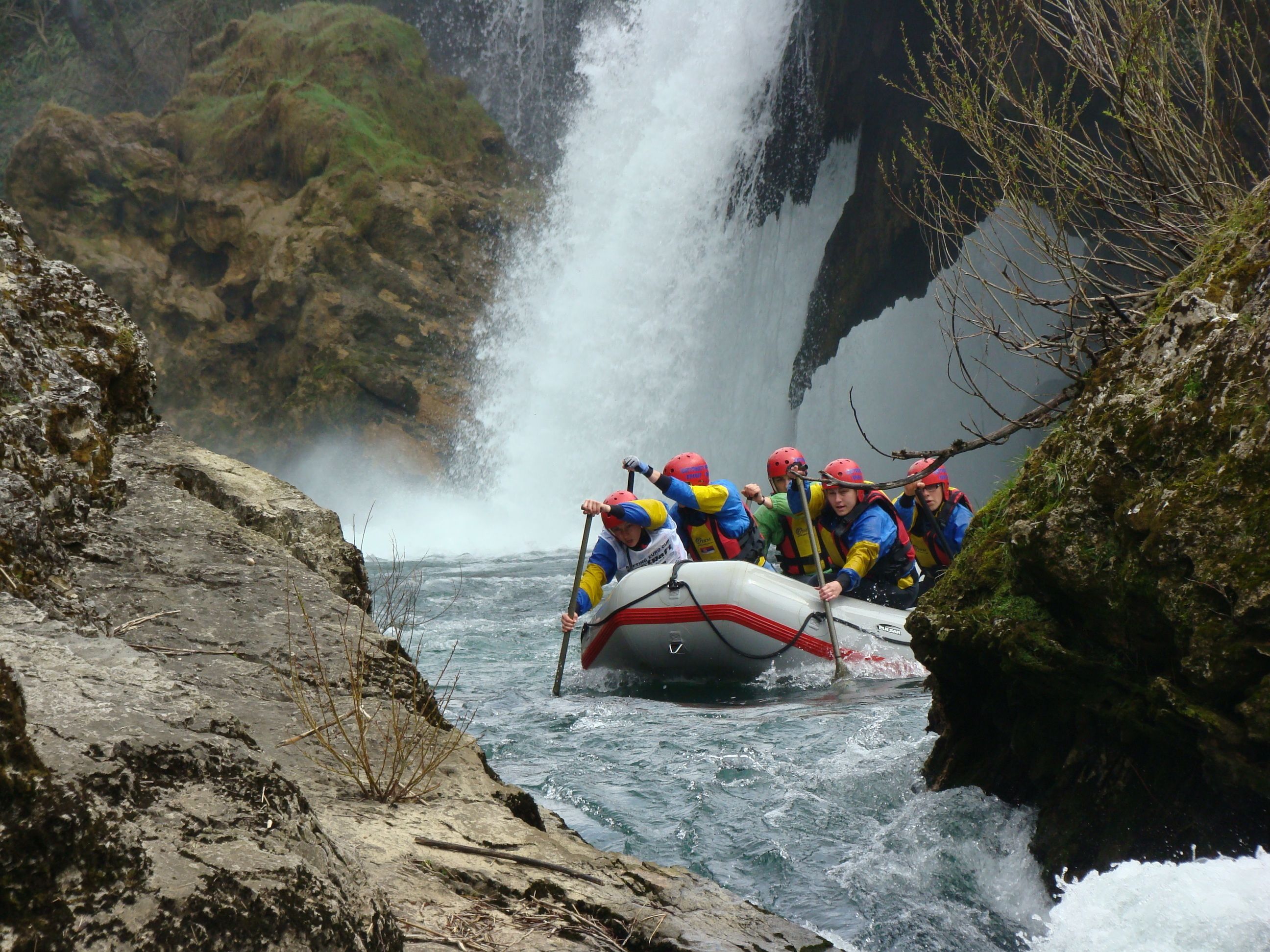 Un groupe de personnes à bord d’un radeau navigue sur une rivière, avec une cascade en arrière-plan.