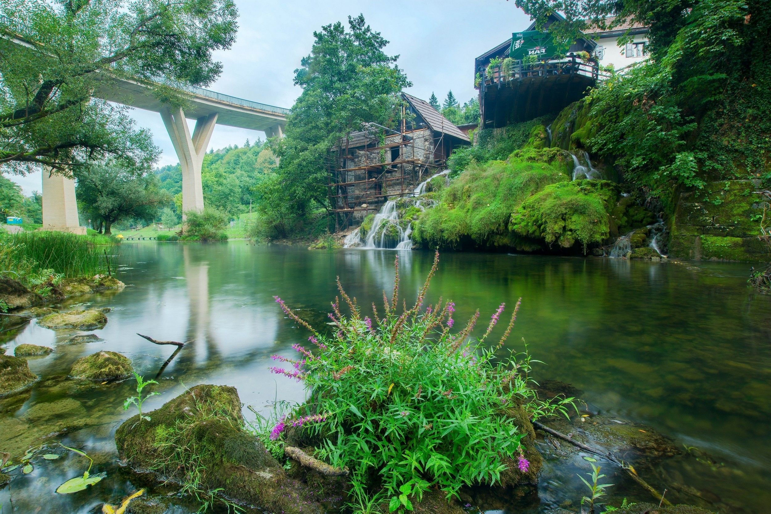 Una cabaña rústica con fachada de madera, una cascada pequeña y un puente al fondo.