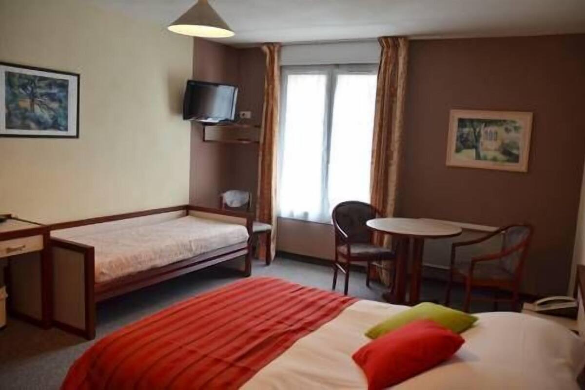 Ein Hotelzimmer mit einem Einzelbett, einem Schreibtisch, einem Sessel, einem Fernseher und einem Fenster mit Vorhängen.