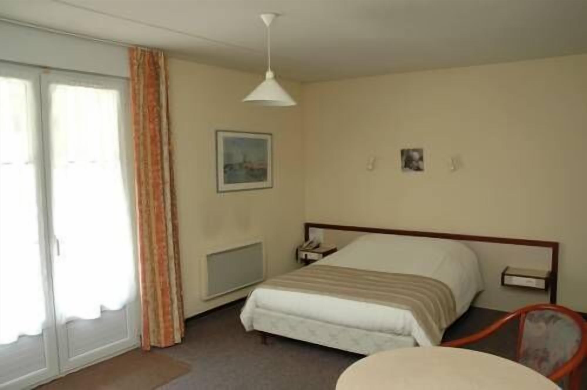 A hotel room with a bed, a bedside table, a chair, a window with curtains, and a ceiling light.