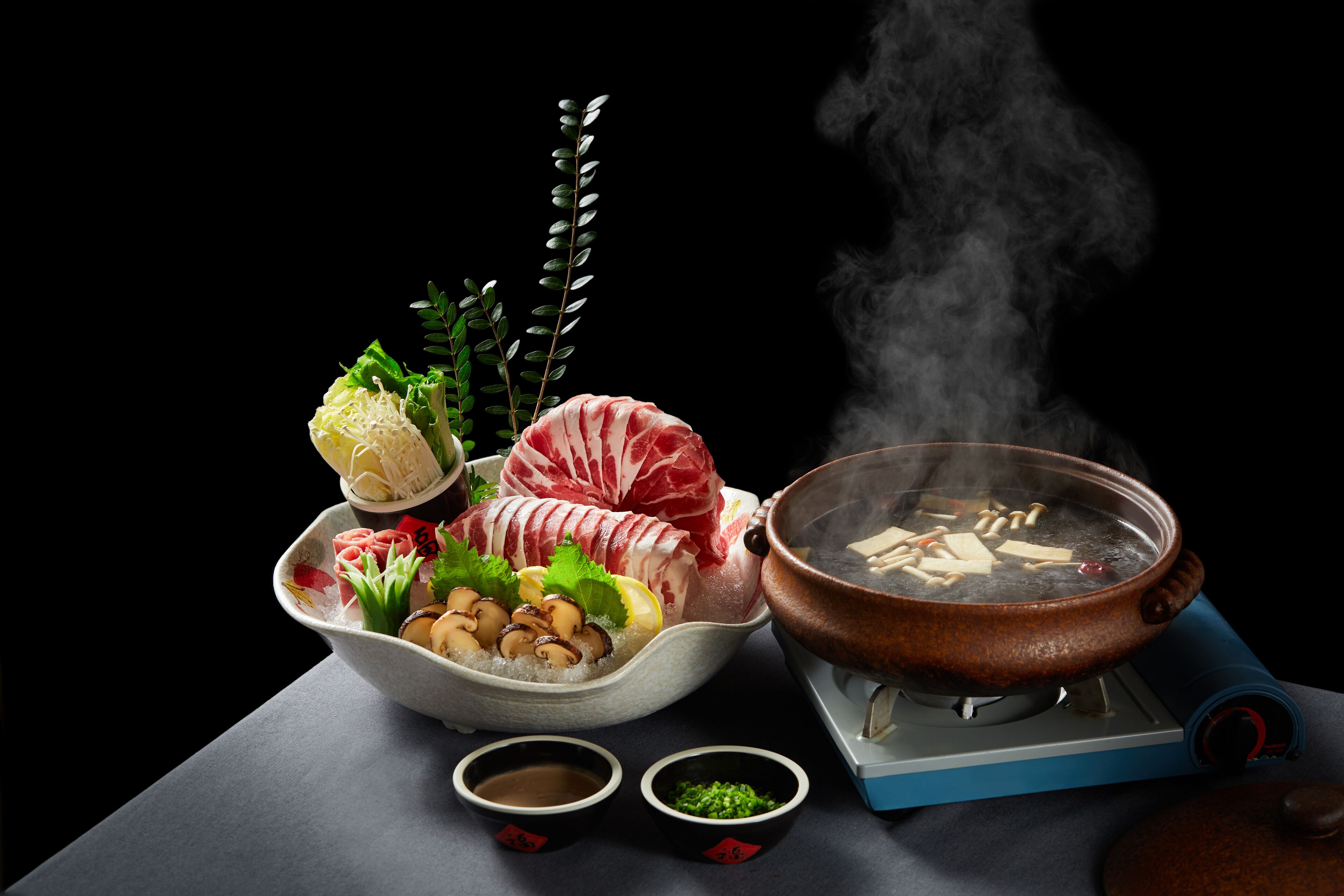 Ein Hot-Pot-Arrangement mit verschiedenen Fleisch- und Gemüsesorten, einem dampfenden Topf und zwei kleinen Saucenschalen.