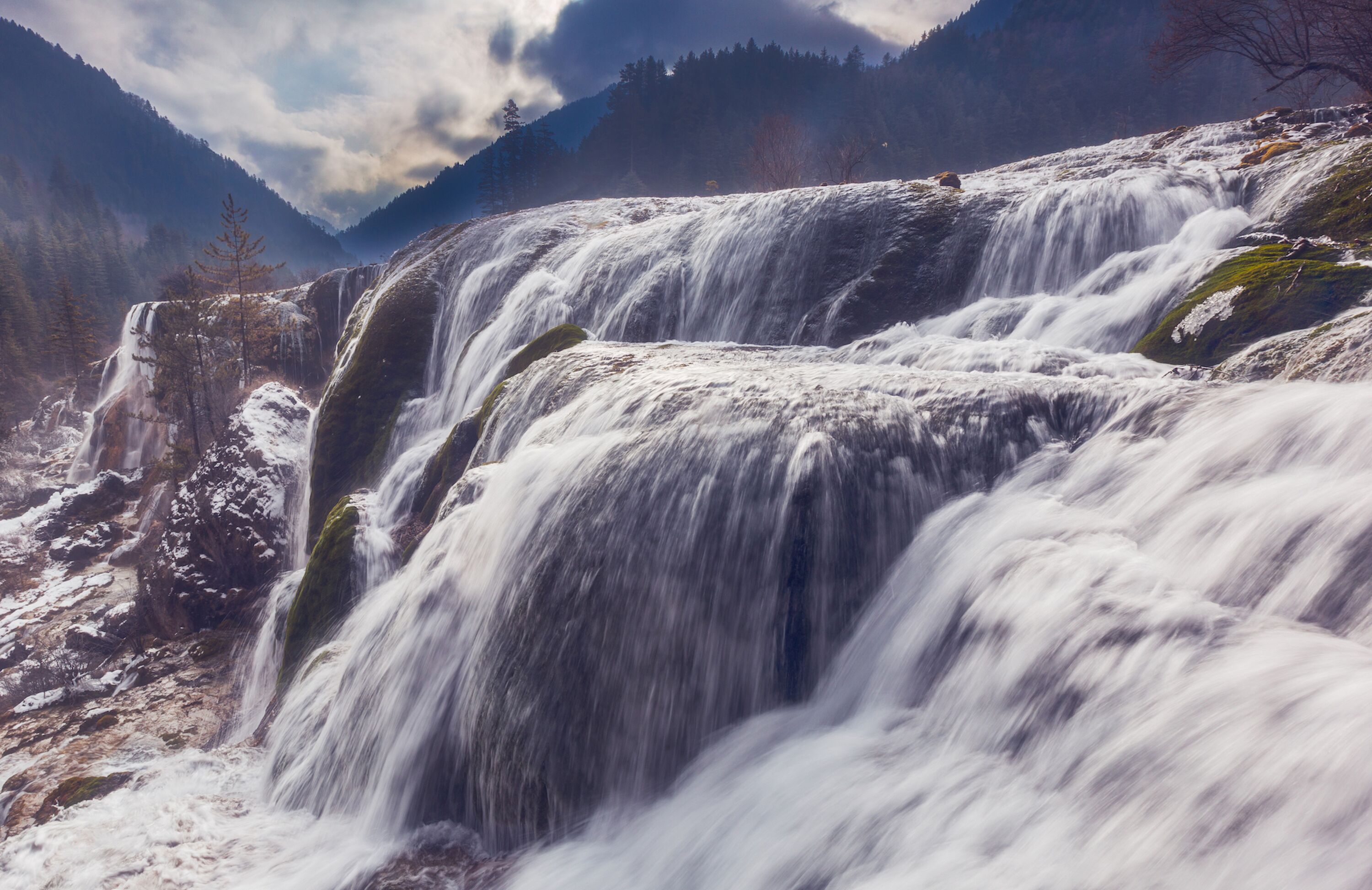 A waterfall cascading over rocks with a forested mountain backdrop.