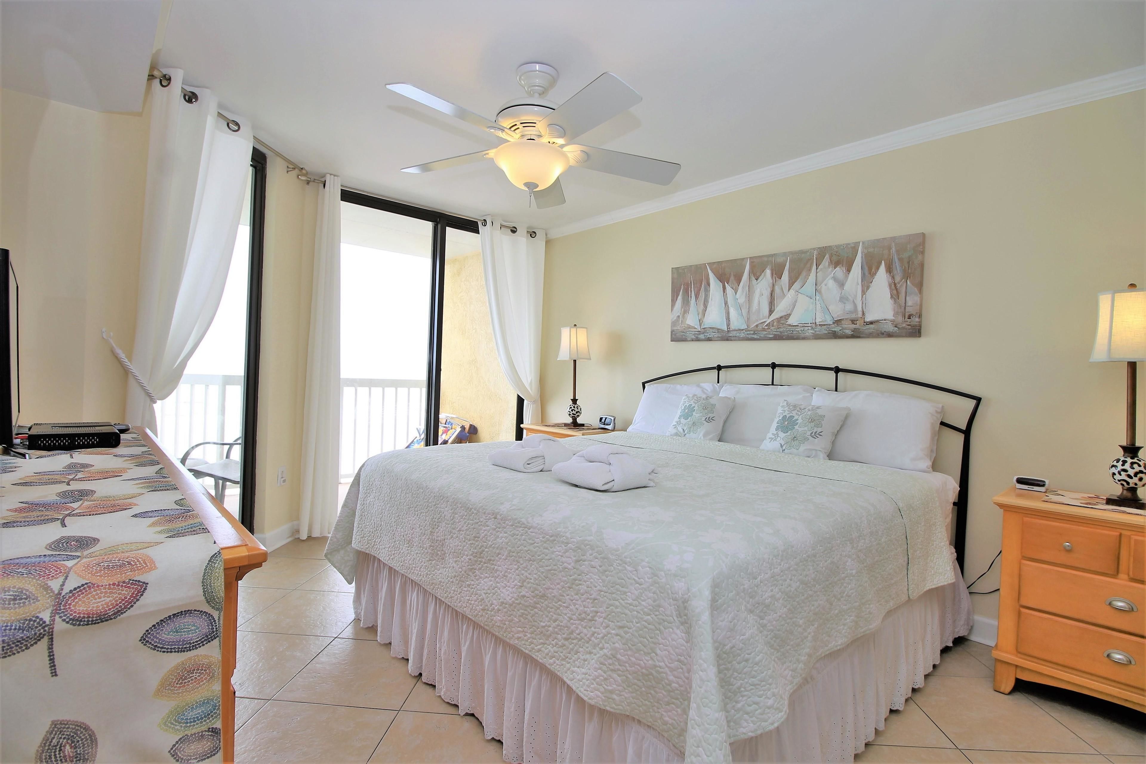 A hotel room with a bed, a nightstand, a table with a tablecloth, a ceiling fan, and a painting on the wall.