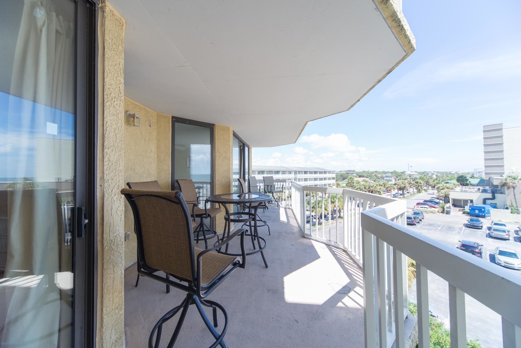 A balcony with a table and chairs, a view of a parking lot, and a building in the background.