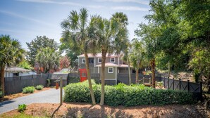 A residential area with a two-story house, a driveway, and a fenced yard with palm trees and shrubs. - Erie The 213 E Hideaway 1 Bedroom Holiday Home By My Ocean Rentals (Folly Beach)