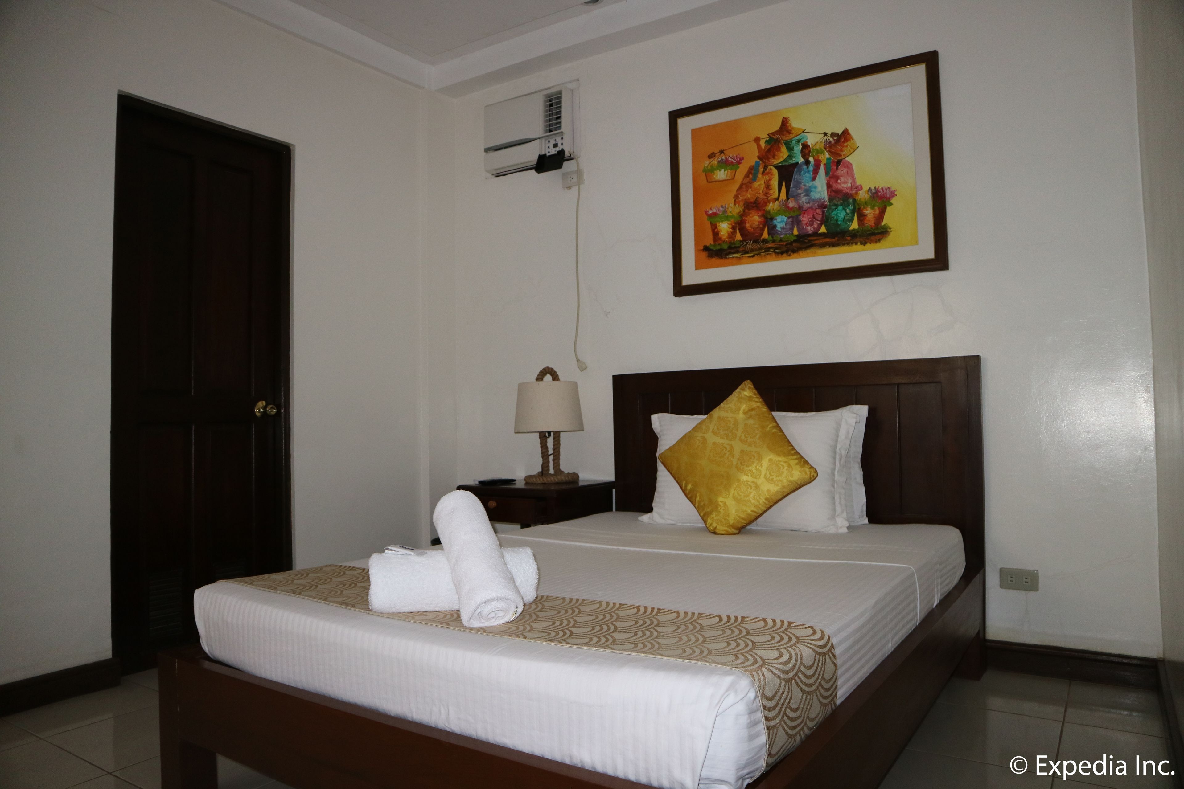 A hotel room with a bed, a nightstand, a lamp, a painting on the wall, and an air conditioning unit.