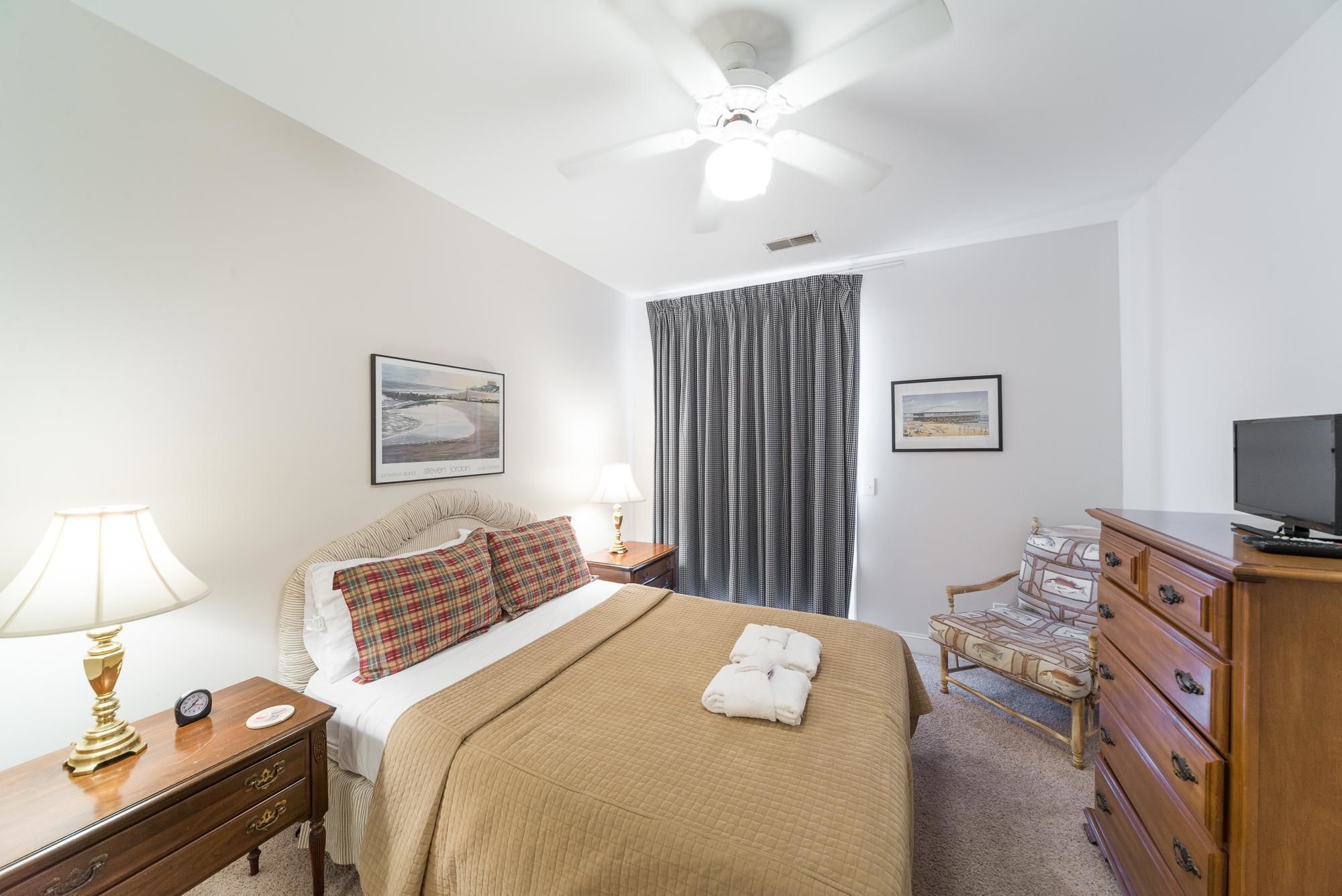A hotel room with a bed, a desk, a chair, a dresser, a ceiling fan, and two framed pictures on the wall.
