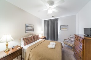 A hotel room with a bed, a desk, a chair, a dresser, a ceiling fan, and two framed pictures on the wall.