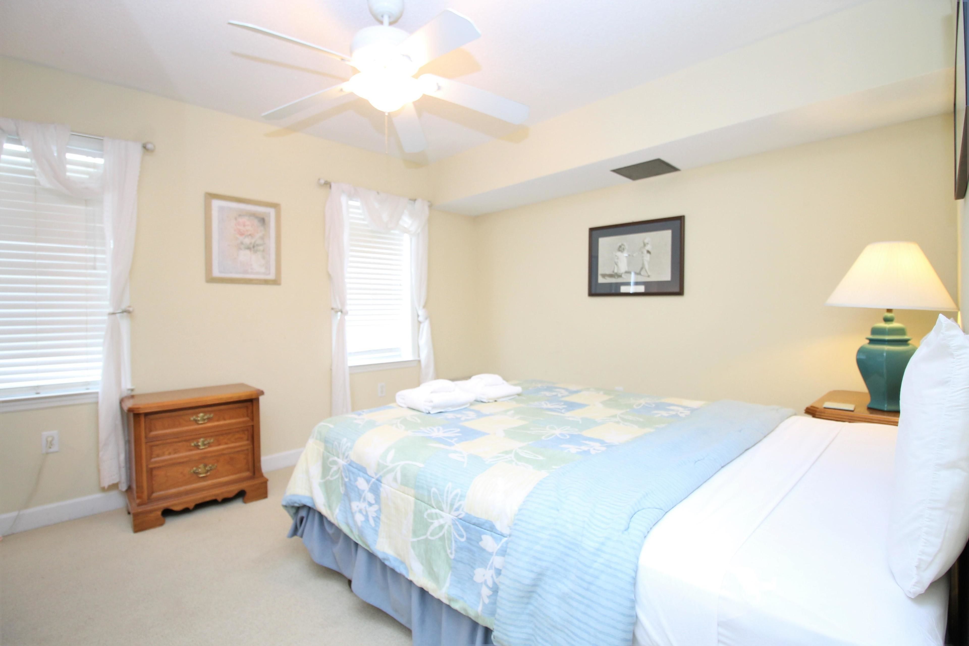 A bedroom with a bed, a nightstand, a dresser, a ceiling fan, and two windows with curtains.