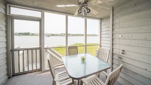 A covered outdoor area with a table and four chairs, a ceiling fan, and large windows offering a view of a body of water and distant buildings. - Mariners Cay 27 2 Bedroom Holiday Home By My Ocean Rentals (Folly Beach)