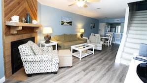 A living room with a fireplace, patterned sofas, a coffee table, and a ceiling fan. - Mariners Cay 27 2 Bedroom Holiday Home By My Ocean Rentals (Folly Beach)