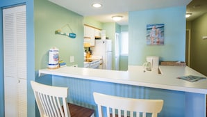 A kitchen with a white countertop, two white chairs, a refrigerator, a coffee maker, and a painting of two children on the wall. - Mariners Cay 27 2 Bedroom Holiday Home By My Ocean Rentals (Folly Beach)
