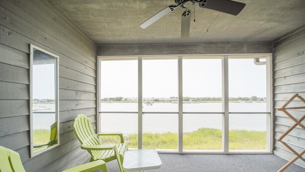 A room with a large window overlooking a body of water, a ceiling fan, and two green chairs with a small table. - Mariners Cay 27 2 Bedroom Holiday Home By My Ocean Rentals (Folly Beach)