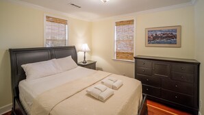 Een opgemaakt bed met een beige deken en twee opgerolde handdoeken, een bedlamp, een donker houten dressoir en een lijst met een afbeelding aan de muur.