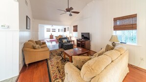 A spacious living room with a large sofa, a coffee table, and a television. There is a ceiling fan, a window with blinds, and a door leading to another room. - E Huron 308 4 Bedroom Holiday Home By My Ocean Rentals (Folly Beach)